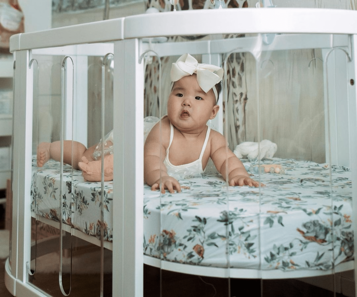 Smiling baby with bow headband in a white playpen on floral bedding, Babyhood GCC