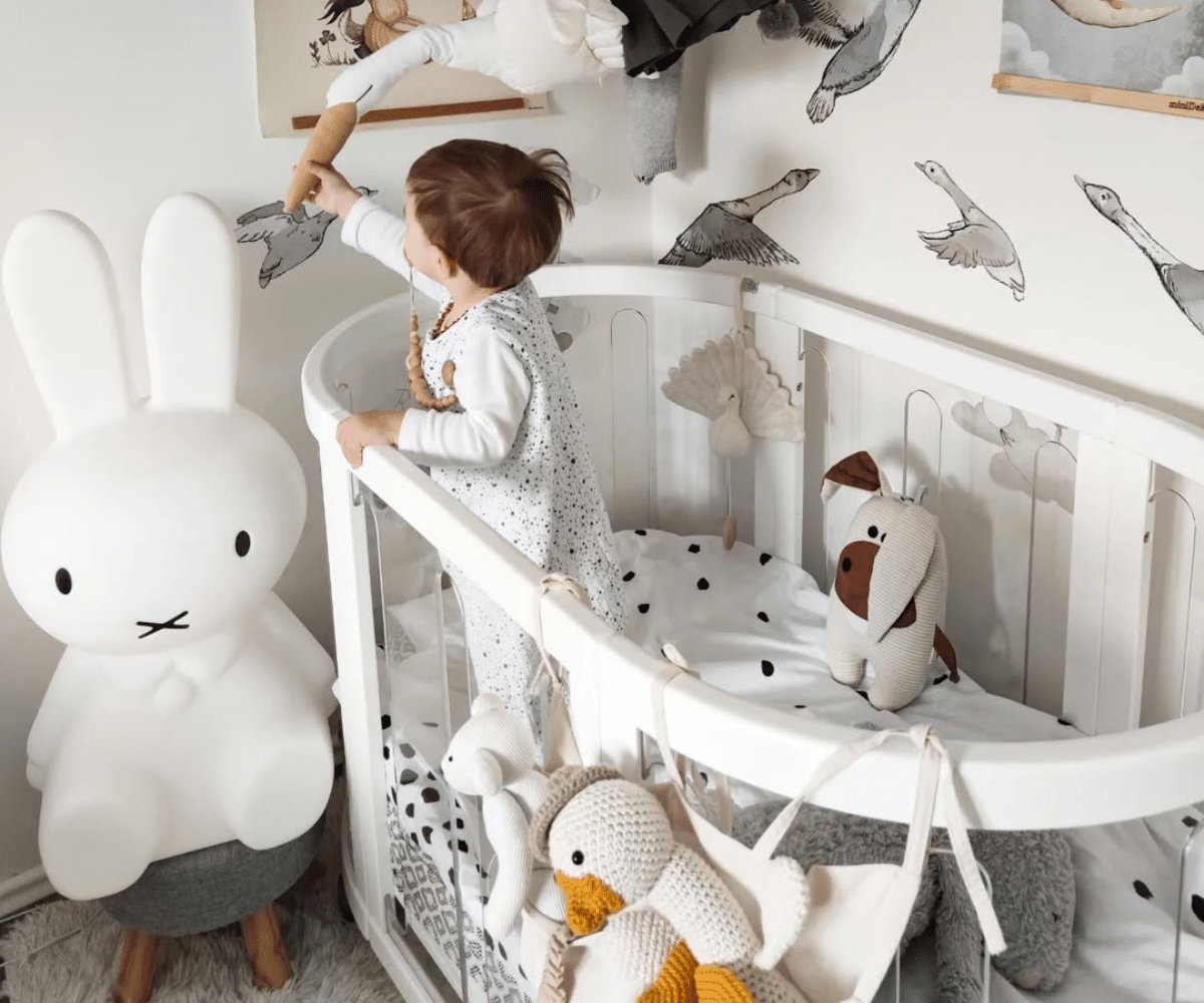 Baby in modern white crib with plush toys, Miffy lamp, and bird wall decals in nursery