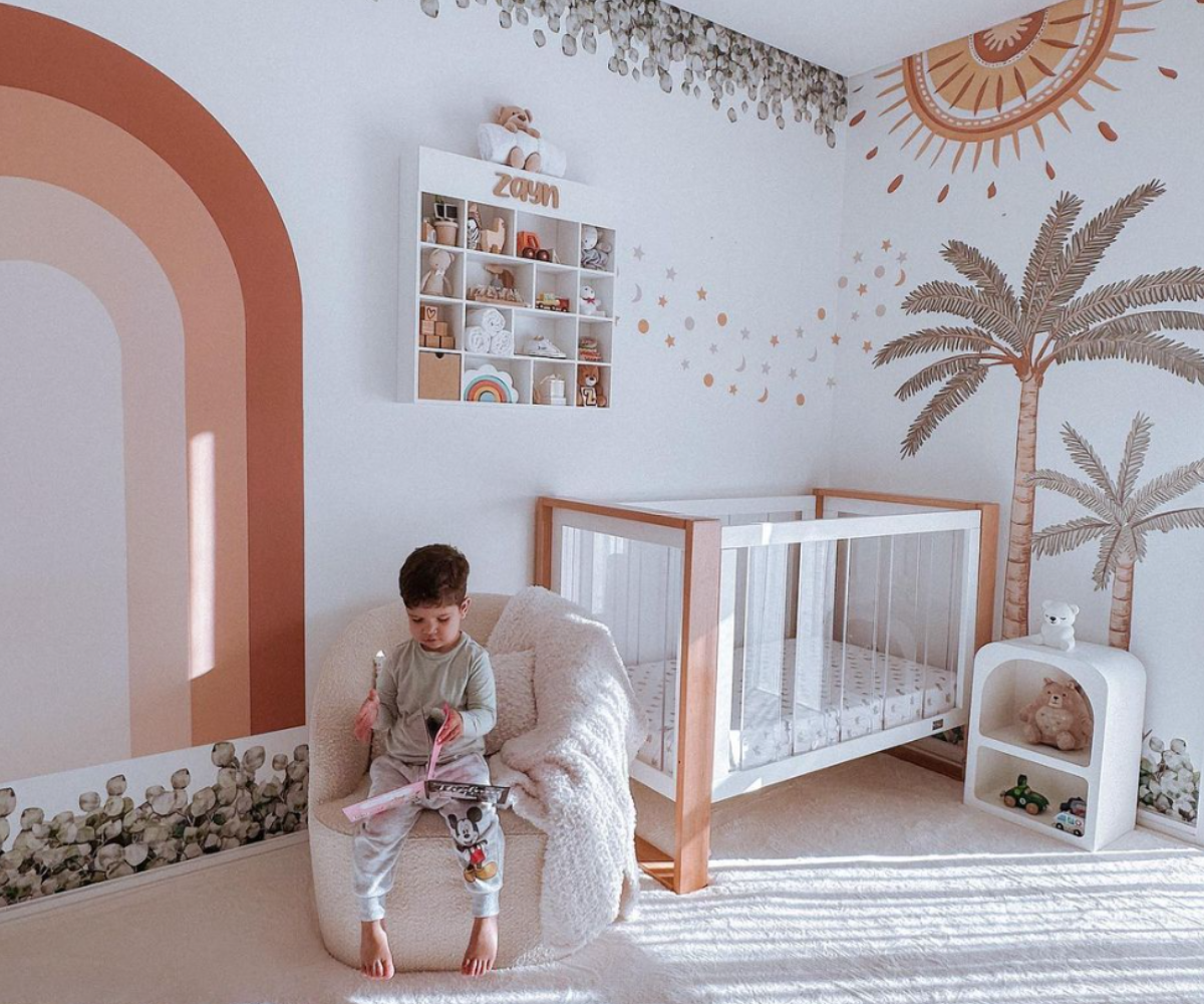 Modern baby nursery with palm tree and rainbow wall art, wooden crib, cozy chair, and toddler