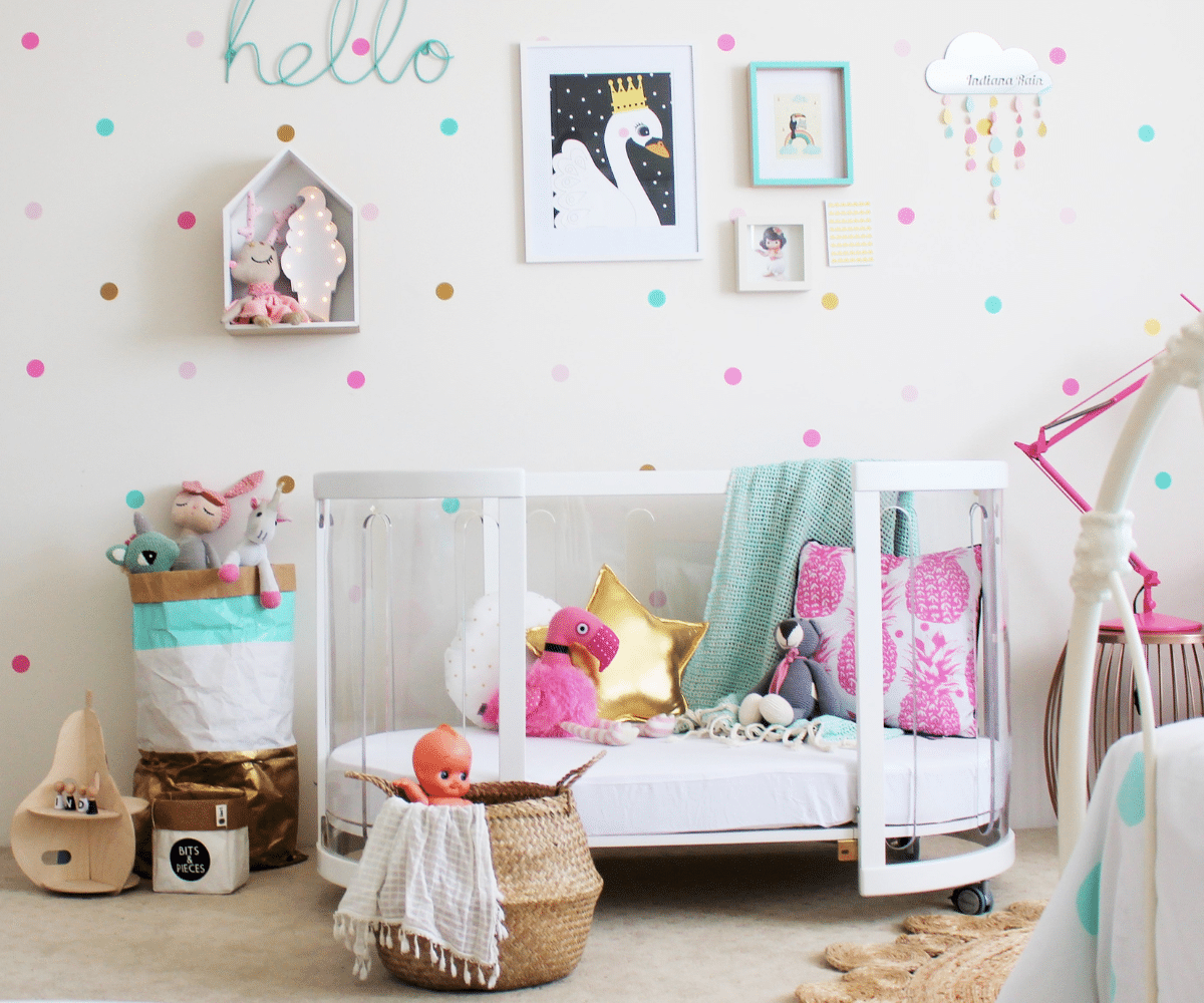 Modern baby nursery with white crib, plush toys, pink and gold decor, and pastel wall dots