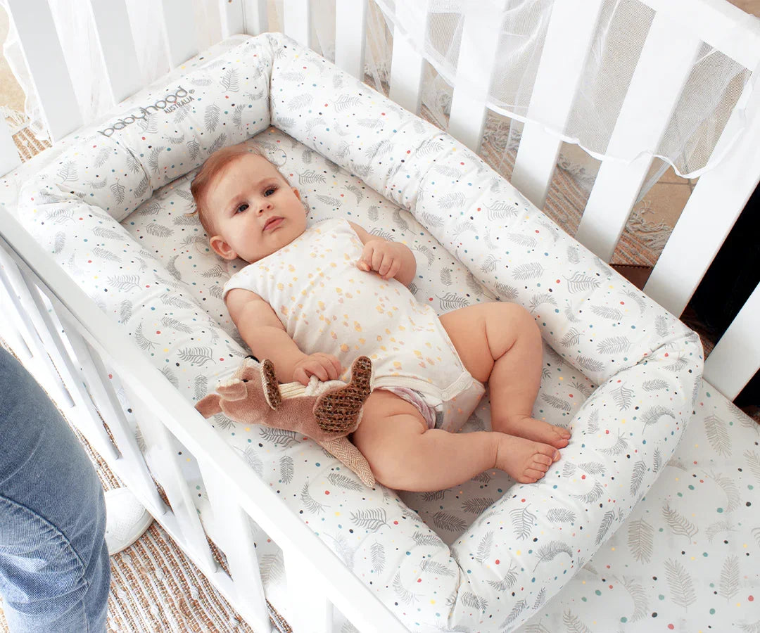 Baby lying in Babyhood cosy crib in white cot with soft patterned bumper and plush toy