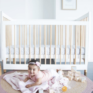 Smiling baby girl on blanket in front of white baby crib, wooden blocks, Babyhood GCC