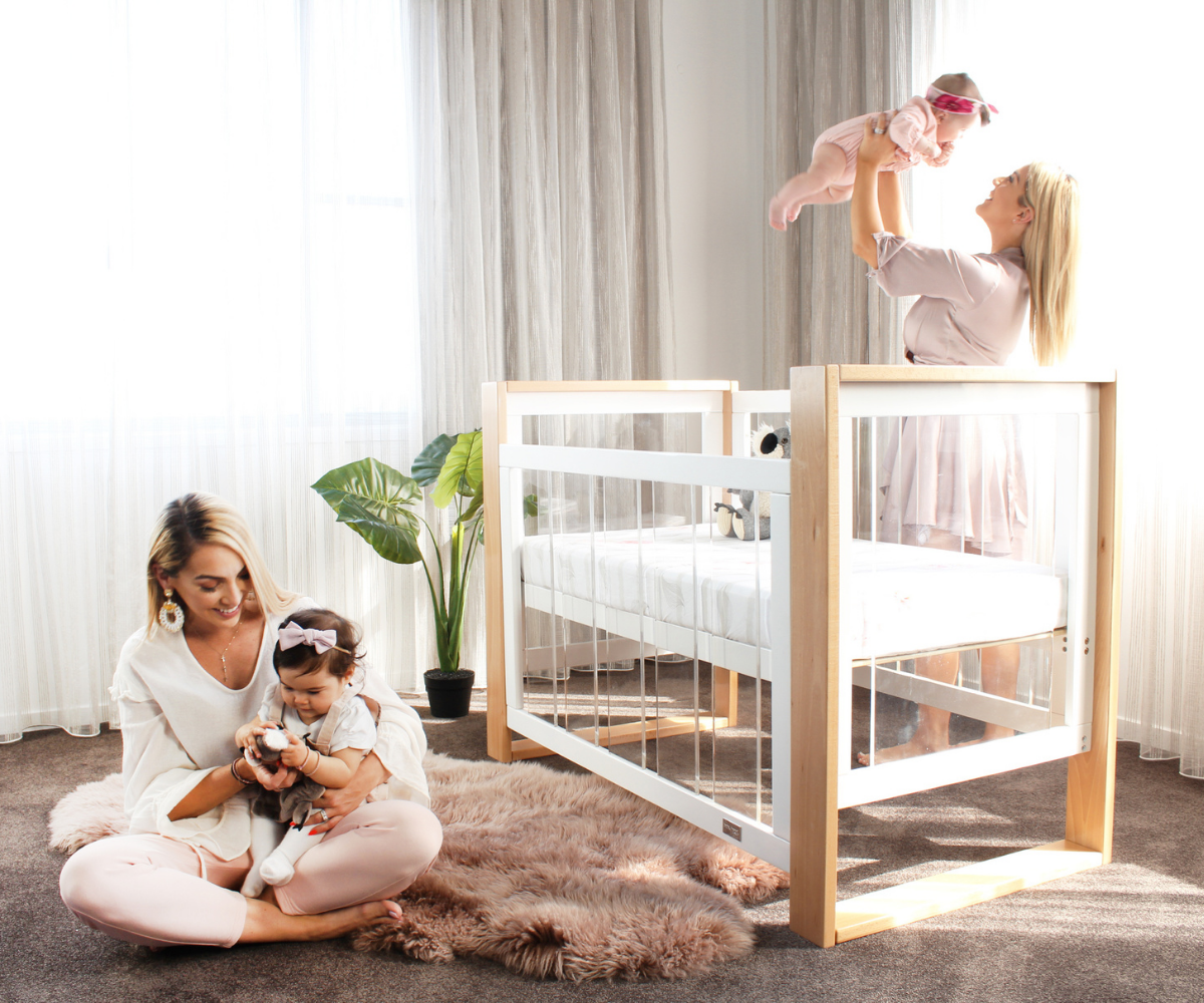 Modern nursery scene with two mothers and babies, clear crib, plush rug, Babyhood GCC