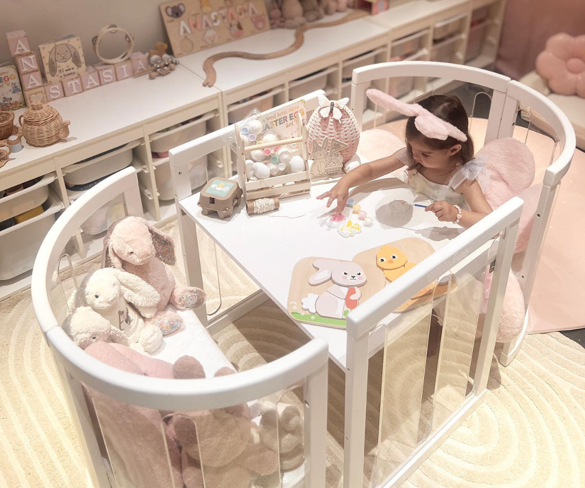 Little girl in pastel nursery playing at white activity table, surrounded by plush toys and Babyhood GCC decor.