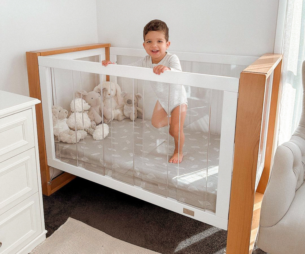 Smiling toddler in a modern Babyhood crib with clear sides and plush toys in a bright nursery