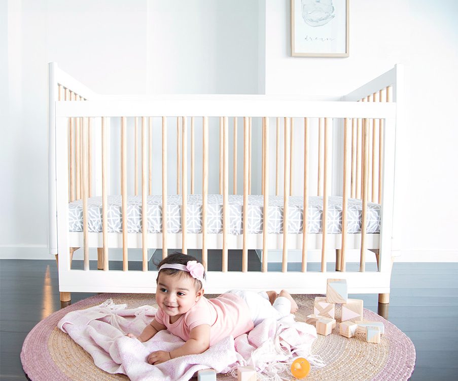 Smiling baby girl on pink mat in front of modern wooden baby cot, styled nursery decor
